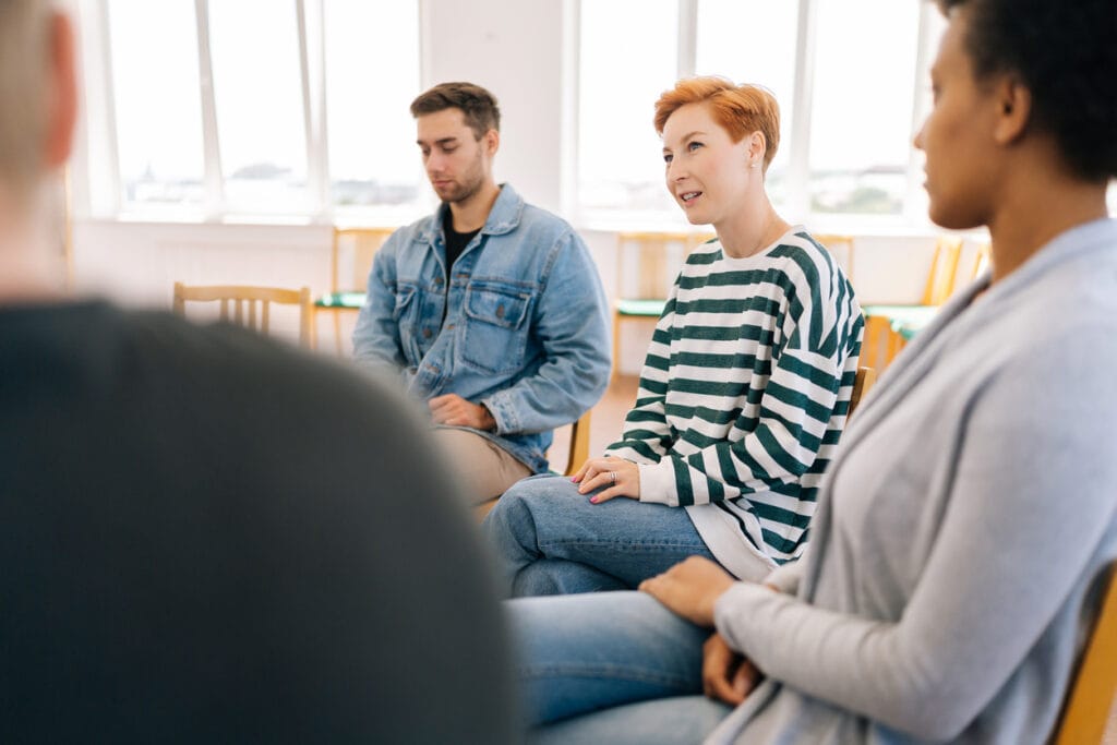 Selective focus of young woman in group psychotherapy session telling about mental problem to patients. Female talking about traumatic experience from past